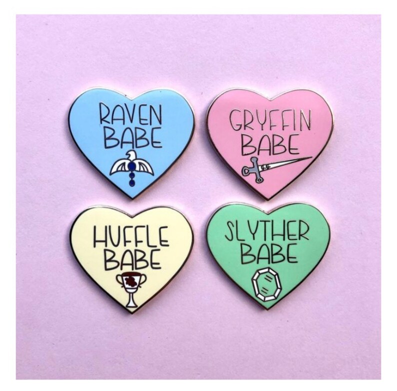 May include: Four heart-shaped enamel pins with different Hogwarts house colors and crests. The pins say "Raven Babe", "Gryffin Babe", "Huffle Babe", and "Slyther Babe".