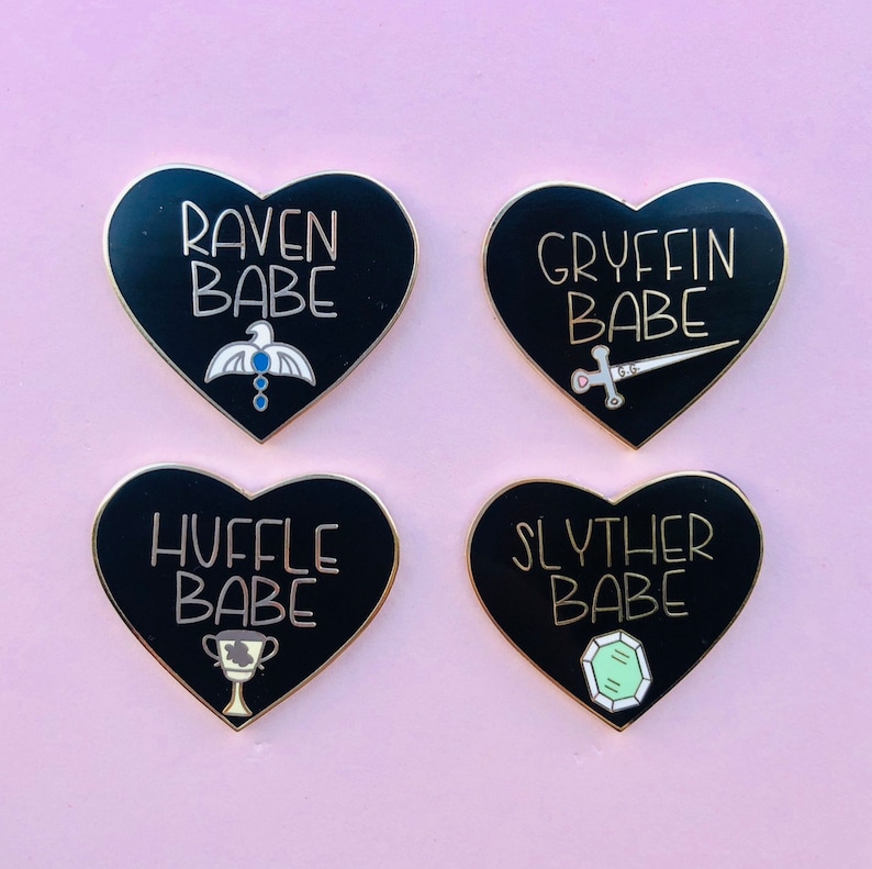 May include: Four black heart-shaped enamel pins with gold accents. Each pin features a different Hogwarts house crest and the text "[House Name] BABE".