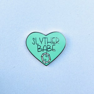 May include: A green heart-shaped enamel pin with the text "Slyther Babe" and a diamond graphic.
