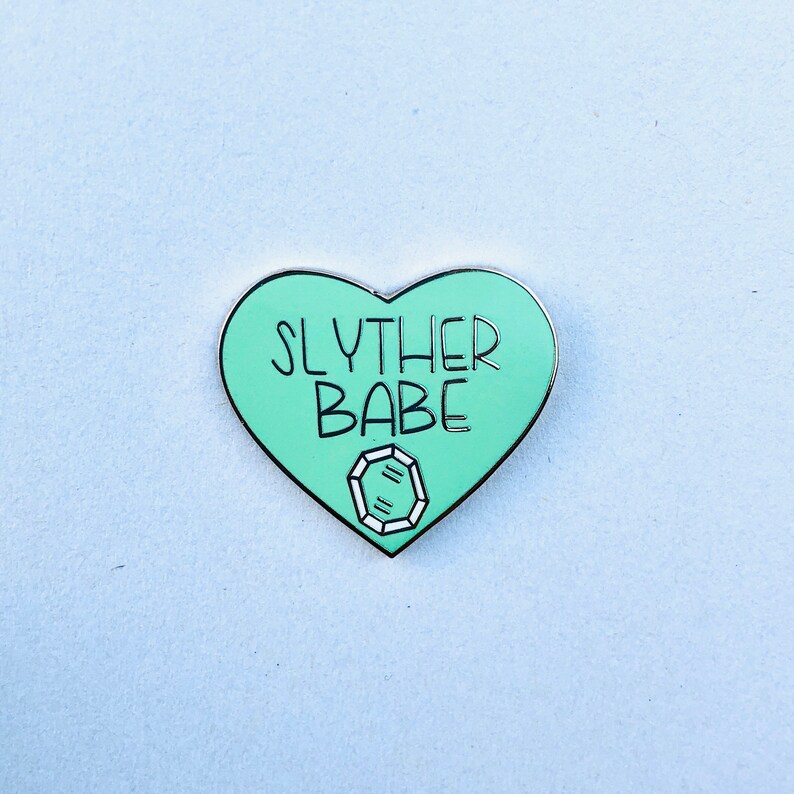 May include: A green heart-shaped enamel pin with the text "Slyther Babe" and a diamond graphic.