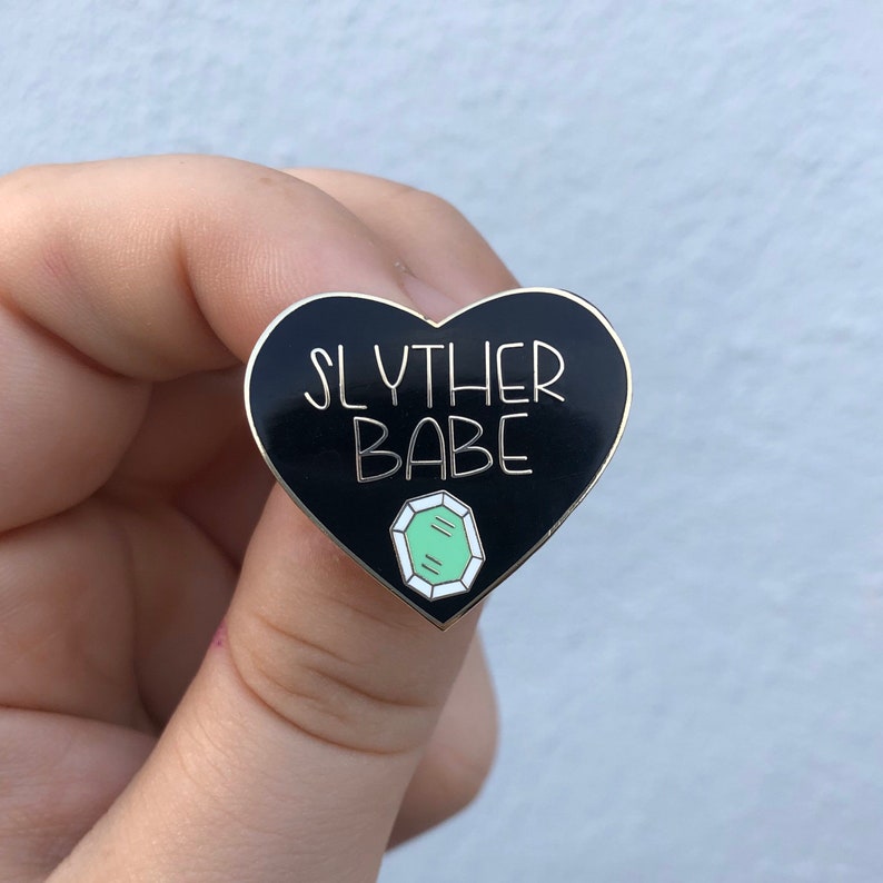 May include: Black heart-shaped enamel pin with the text "Slyther Babe" in white lettering and a green gemstone graphic.