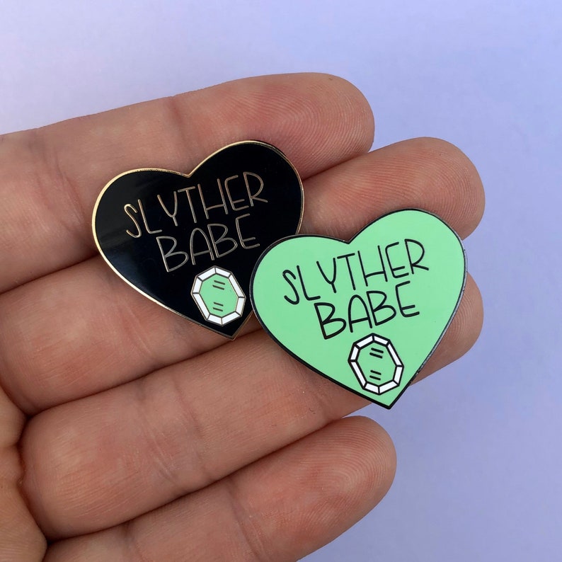 May include: Two heart-shaped enamel pins with the text "Slyther Babe" in gold lettering. One pin is black with a green gem, and the other is green with a white gem.