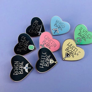 May include: Set of nine heart-shaped enamel pins featuring the Hogwarts houses. Each pin has a different house crest and the text "[House Name] Babe". The pins are black, pink, blue, yellow, and green.