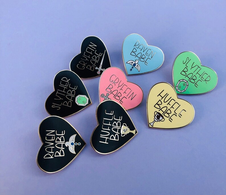 May include: Set of nine heart-shaped enamel pins featuring the Hogwarts houses. Each pin has a different house crest and the text "[House Name] Babe". The pins are black, pink, blue, yellow, and green.