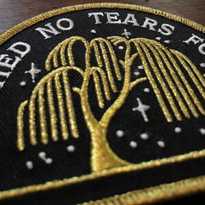 Op de afbeelding: Een zwarte geborduurde patch met gouden draad die een treurwilg voorstelt met de tekst "I'll Shed No Tears For" boven de boom en "You" onder de boom.