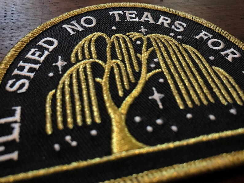 Op de afbeelding: Een zwarte geborduurde patch met gouden draad die een treurwilg voorstelt met de tekst "I'll Shed No Tears For" boven de boom en "You" onder de boom.