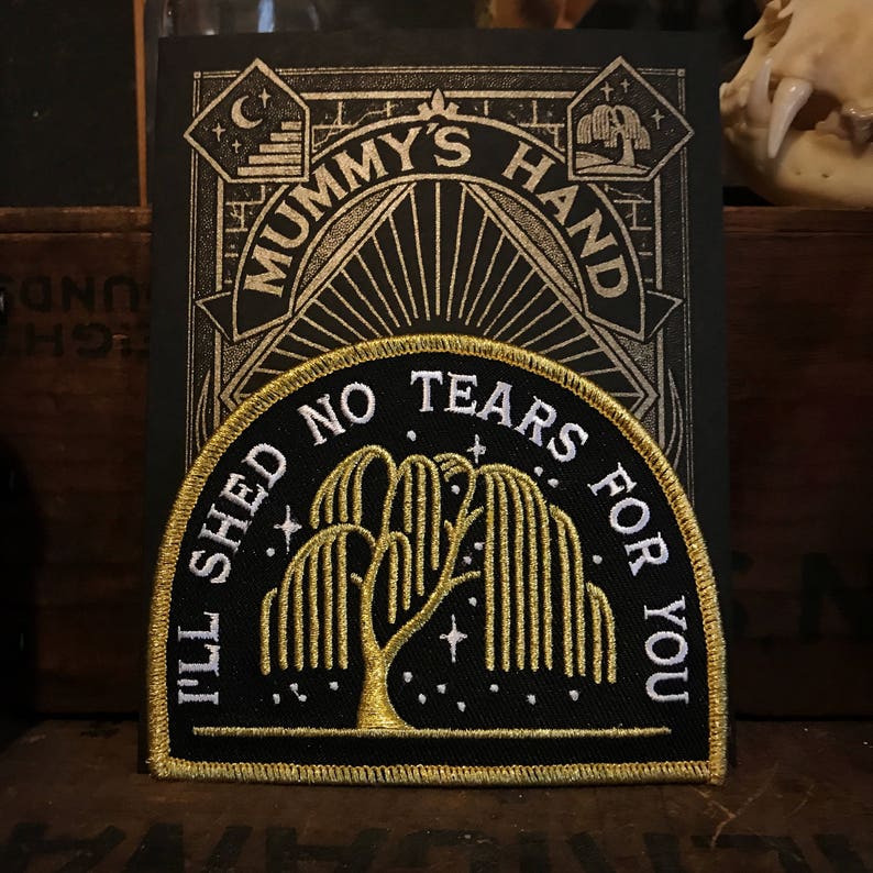 Op de afbeelding: Een geborduurde zwarte en gouden patch met een treurwilg-ontwerp. De tekst luidt "I'll Shed No Tears For You".