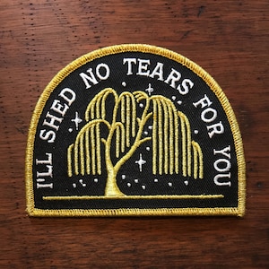 Op de afbeelding: Een zwarte geborduurde patch met een gouden rand. De patch heeft een treurwilg met witte sterren en de tekst "I'll shed no tears for you".