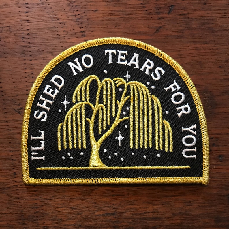 Op de afbeelding: Een zwarte geborduurde patch met een gouden rand. De patch heeft een treurwilg met witte sterren en de tekst "I'll shed no tears for you".