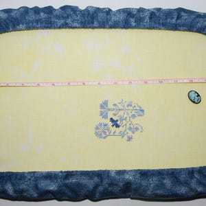 May include: A blue and yellow embroidery hoop with a floral design stitched in purple, blue, and white thread. A small blue button is in the top right corner of the hoop.