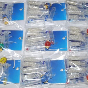 May include: A set of 10 small clear plastic tubes filled with white glitter. The tubes are arranged in three rows of three, with one tube in the bottom row. Each tube has a small, colorful charm attached to the top.