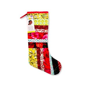 May include: A red, yellow, and black patchwork Christmas stocking with a floral pattern. The stocking has a tag attached with the text "Women of Color" and a picture of a woman.