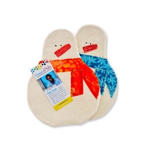 May include: Two white fabric snowman-shaped pot holders with red and blue fabric accents. The pot holders have red and blue fabric scarves and black embroidered eyes and mouths. A small card with the text "Women of  Mild" is visible in the image.