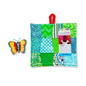 May include: A colorful patchwork pot holder with a green, blue, and white color scheme. The pot holder has a red loop for hanging. A small card with the text "Women of Milot Entrepreneurial Network" and a photo of a woman is attached to the pot holder. The text "Zoulos" is also on the card.  The text "#GIVINGBACK" is at the bottom of the card.
