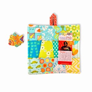 May include: A colorful patchwork hot pad with a hanging loop. The hot pad is made of various fabrics in shades of blue, green, yellow, orange, and white. A small tag with the text "Women of Milot ENTREPRENEURIAL NETWORK" is attached to the hot pad. The tag also includes a photo of a woman and a short description of her story.