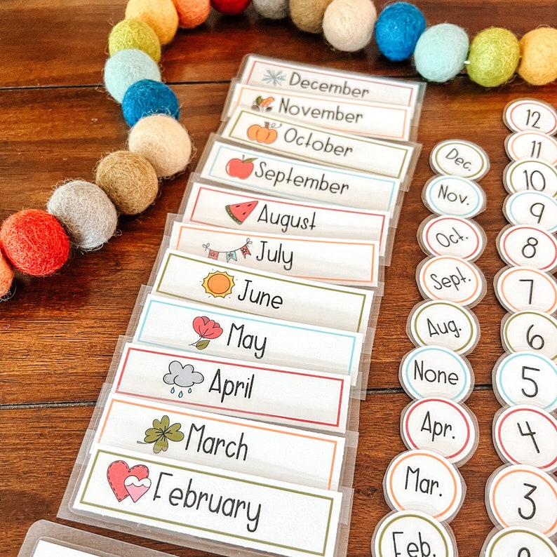 Puede incluir: Un conjunto de 12 tarjetas coloridas con ilustraciones y los nombres de cada mes del a&ntilde;o. Las tarjetas est&aacute;n dispuestas en una pila vertical, con diciembre en la parte superior y febrero en la parte inferior. Las tarjetas est&aacute;n sobre una superficie de madera con una guirnalda de bolas de colores.