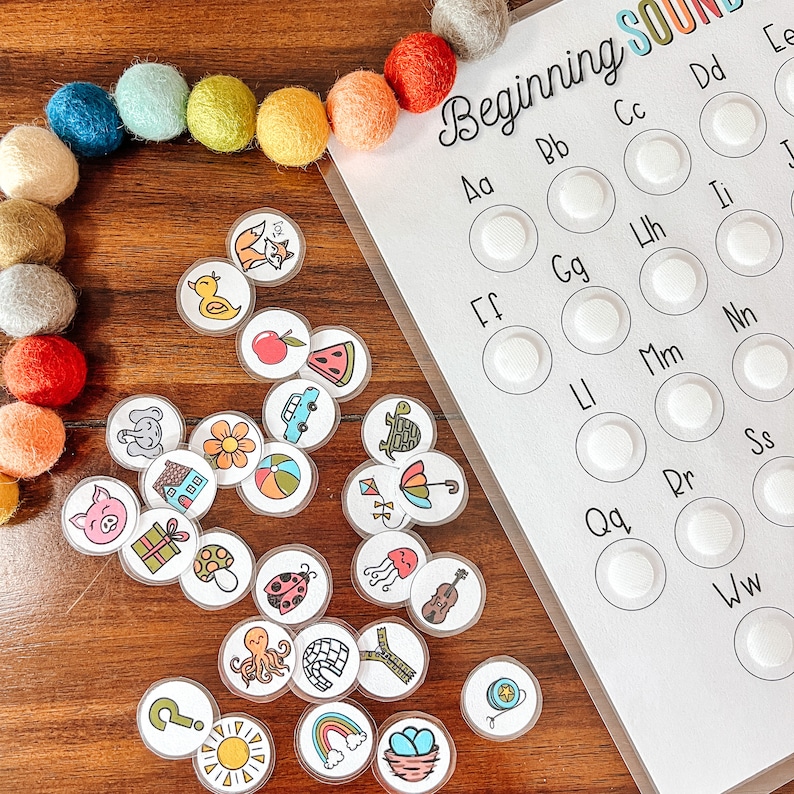 May include: A white board with the words "Beginning Sounds" written in black marker. The board has a grid of circles with letters written in black marker. There are 20 circles with letters from A to W. There are colorful circles with pictures of various objects on them, such as a turtle, a pig, a flower, a watermelon, a car, a ladybug, a mushroom, a sun, a rainbow, and an octopus.