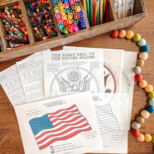 May include: A wooden box filled with colourful crayons and markers sits on a wooden surface, with colouring pages featuring the American flag and the Great Seal of the United States. A felt ball garland adds a decorative touch.