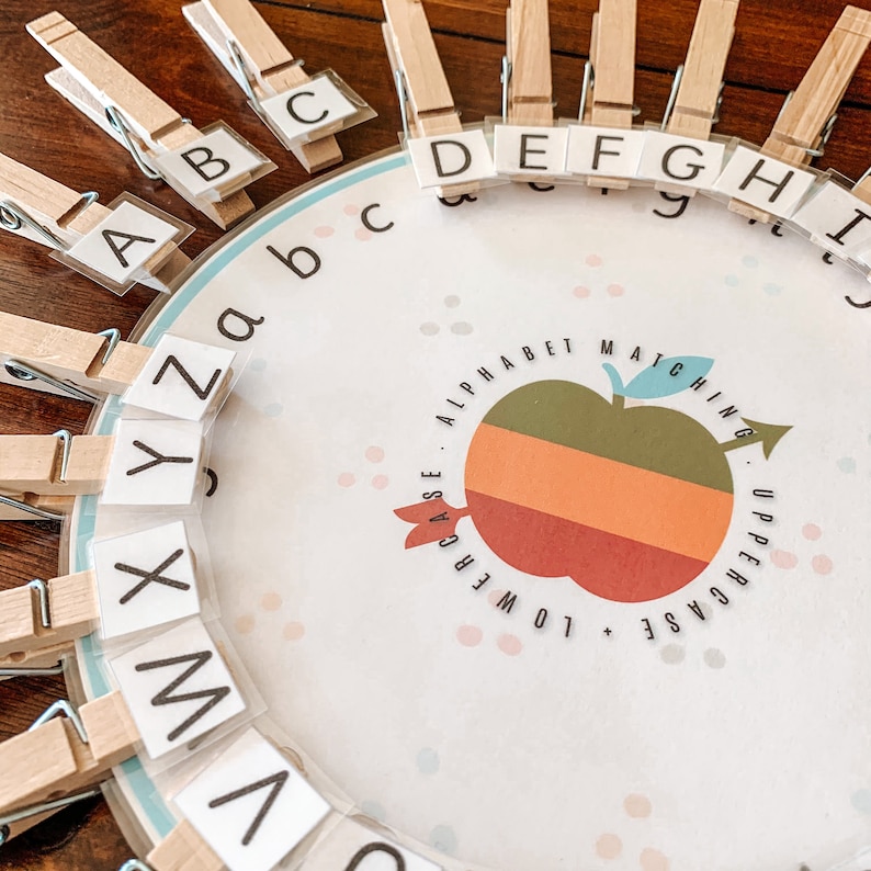 May include: A white circular board with a colorful apple graphic in the center. The board has a blue border and is covered in white dots. The board is labeled "Alphabet Matching: Uppercase + Lowercase".  Wooden clothespins with letters of the alphabet are attached to the board.