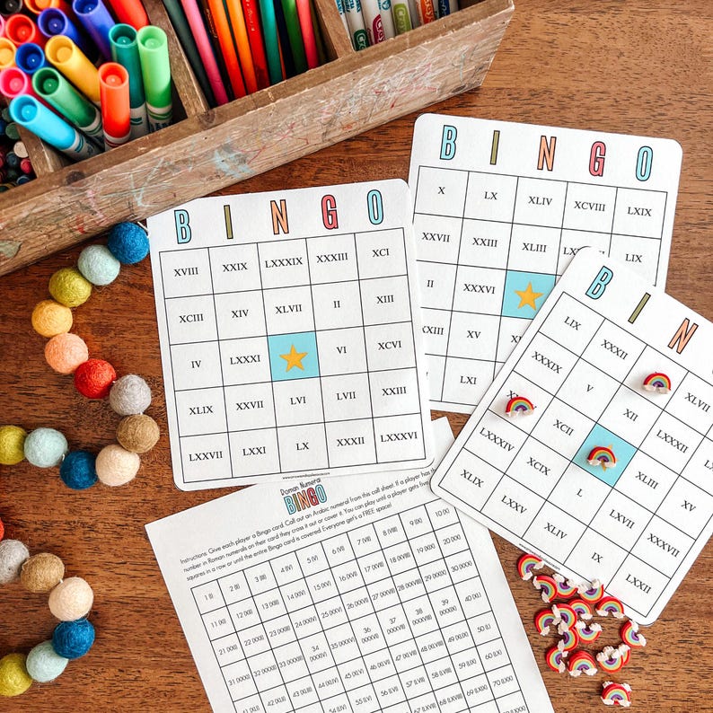 May include: Three colorful bingo cards with Roman numerals and a star in the center. The cards are on a wooden table with a pile of rainbow-colored felt balls and a sheet of paper with a bingo number chart.