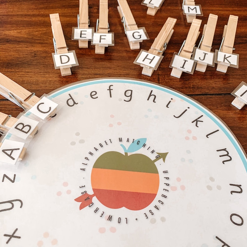 Puede incluir: Un juego de asociaci&oacute;n de alfabeto colorido con un dise&ntilde;o de manzana roja, naranja y verde. El juego incluye un tablero circular con el alfabeto impreso alrededor del borde y fichas de letras individuales con pinzas para la ropa.