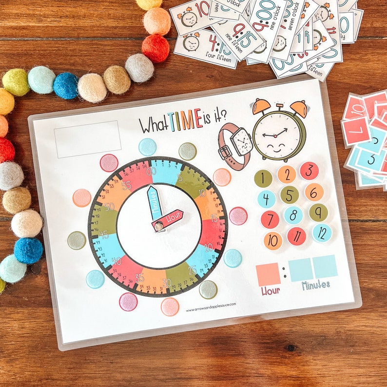 Peut inclure: Un cadran d'horloge color&eacute; avec des aiguilles mobiles pour apprendre aux enfants &agrave; dire l'heure. Le cadran de l'horloge est entour&eacute; de cercles color&eacute;s avec les chiffres de 1 &agrave; 12 et des incr&eacute;ments de 5 minutes. Le cadran de l'horloge a &eacute;galement une aiguille des heures et une aiguille des minutes s&eacute;par&eacute;es. Le texte "What TIME is it?" est en haut du cadran de l'horloge.