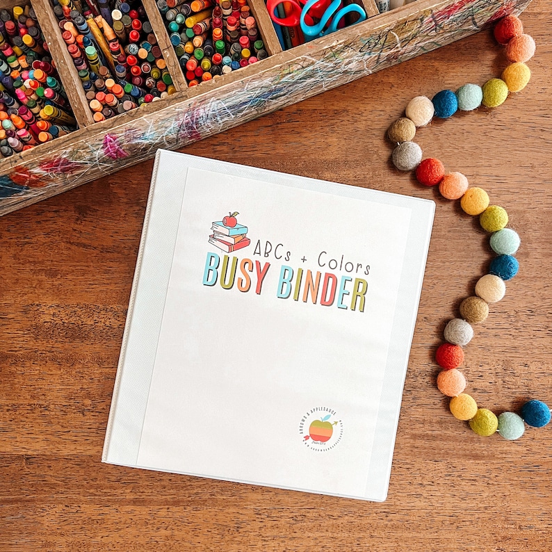 Puede incluir: Una carpeta blanca con el texto "ABCs + Colors Busy Binder" en la parte delantera. La carpeta est&aacute; sobre una mesa de madera con una cadena de pompones de colores.