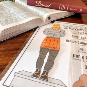 May include: A printable paper doll of a man with a gold crown, orange skirt, and gray legs. The paper doll is standing on a platform. The text on the paper doll says "Nebuchadnezzar's Dream".