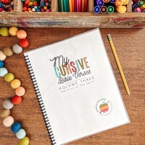 Puede incluir: Un cuaderno blanco con un título colorido "My Cursive Bible Verses Volume Three The Fruit of the Spirit" y un pequeño logotipo de manzana. El cuaderno está abierto y tiene una encuadernación en espiral.