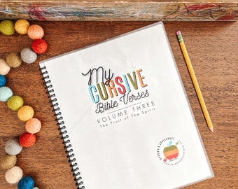 May include: A white notebook with a colorful title "My Cursive Bible Verses Volume Three The Fruit of the Spirit" and a small apple logo. The notebook is open and has a spiral binding.
