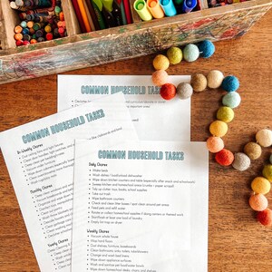 May include: Two printable checklists for common household tasks, with categories for daily, weekly, and monthly chores. The checklists are printed on white paper and are lying on a wooden table with a colorful string of beads.