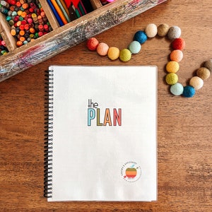 May include: A white spiral bound notebook with the text "The PLAN" in colourful letters. The notebook has a small apple logo with the text "Around the Apple Orchard" on the bottom right corner.