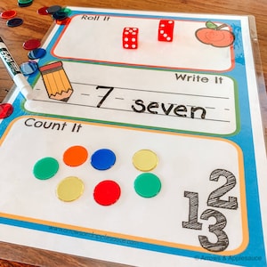 K&ouml;nnte beinhalten: Ein wei&szlig;es laminiertes Lernblatt mit den Worten "Roll It", "Write It" und "Count It" darauf gedruckt. Das Blatt enth&auml;lt einen roten W&uuml;rfel, einen Bleistift und einen roten Apfel mit einem l&auml;chelnden Gesicht. Es gibt sieben bunte Kreise und die Zahlen 12 und 13 auf dem Blatt.