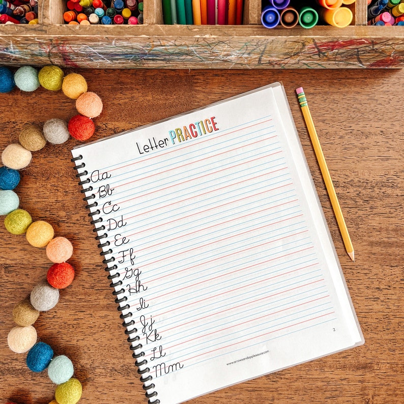 May include: A spiral bound notebook with lined pages for practicing cursive handwriting. The cover has the words "Letter Practice" in colorful letters. The notebook is on a wooden table with a yellow pencil and a colorful garland of felt balls.