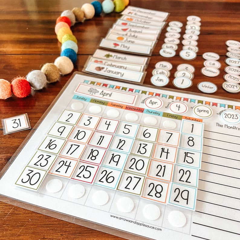 Puede incluir: Un calendario blanco con el mes de abril resaltado. El calendario tiene un dise&ntilde;o colorido con cuadrados de diferentes colores para cada d&iacute;a del mes. El calendario est&aacute; sobre una mesa de madera con una cuerda de pompones coloridos.