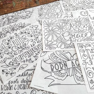 May include: A collection of black and white coloring pages with floral and inspirational designs. The pages feature phrases like "His Grace is Sufficient" and "Born Again," along with floral and butterfly illustrations. The pages are scattered on a wooden surface.