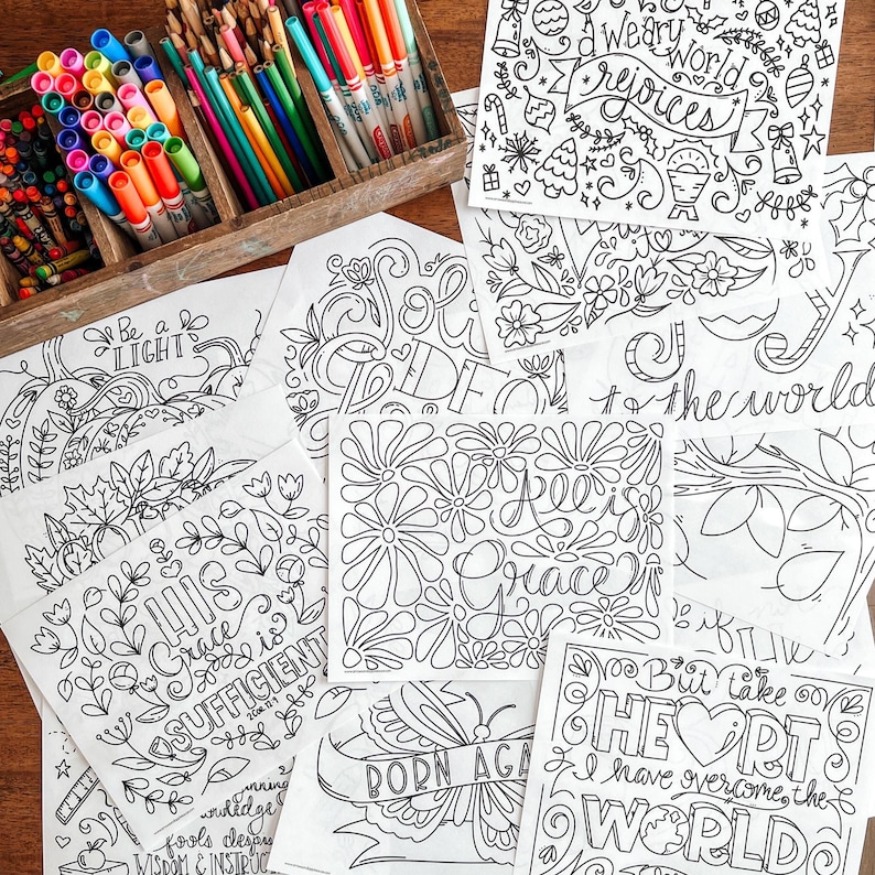 May include: A collection of coloring pages with intricate floral and inspirational designs, alongside a wooden box filled with colorful markers. The pages feature phrases like "Grace is Sufficient" and "Rejoices". Perfect for creative expression.