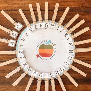 May include: An alphabet matching game with wooden pegs arranged around a circular design. The game features uppercase and lowercase letters and a central apple image. The pegs are attached to the letters.