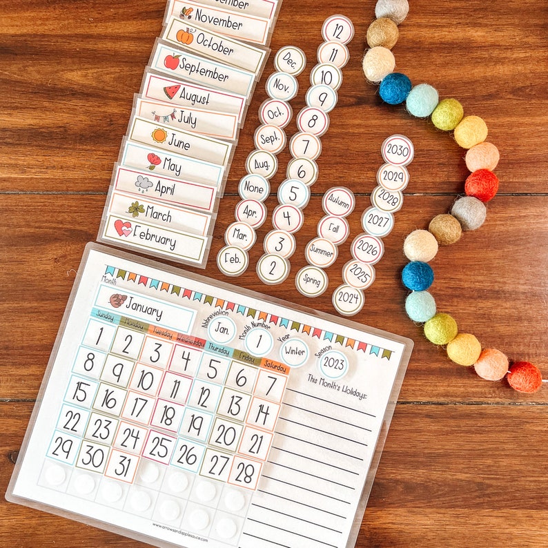 Puede incluir: Un calendario blanco con una cuadr&iacute;cula de calendario de enero de 2023, una secci&oacute;n para los d&iacute;as festivos del mes y una secci&oacute;n para las estaciones del a&ntilde;o. El calendario est&aacute; rodeado de bolas de fieltro de colores y un juego de tarjetas de meses y n&uacute;meros.