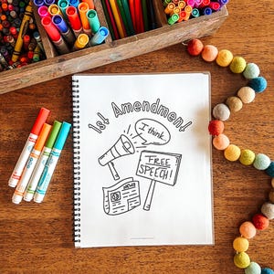 Puede incluir: Un cuaderno de espiral con una ilustraci&oacute;n en blanco y negro sobre la 1&ordf; Enmienda y la libertad de expresi&oacute;n. La imagen incluye un meg&aacute;fono, un peri&oacute;dico y un letrero que dice "&iexcl;Libertad de expresi&oacute;n!" Rotuladores y l&aacute;pices de colores rodean el cuaderno.