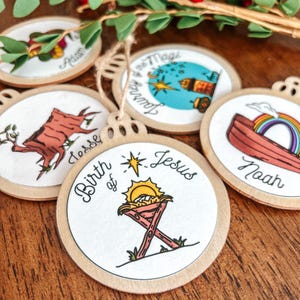 May include: Set of six wooden Christmas ornaments with illustrations and text. The ornaments feature illustrations of a manger, a tree stump, a boat, a star, and a town. The text on the ornaments includes "Birth of Jesus", "Jesse", "Noah", "Journey of the Magi", and "Adam".