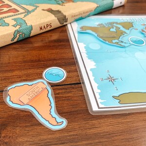 May include: A set of colorful, laminated world map puzzle pieces with a compass, an orange South America continent piece, and a blue "Pacific" circle piece. The pieces are on a wooden surface.