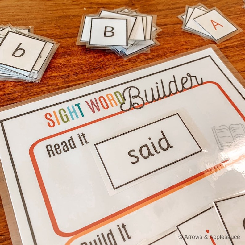 May include: A white card with the words "Sight Word Builder" in colorful letters. The card has a section that says "Read it" and a section that says "Build it". The word "said" is written on a white card and placed in the "Read it" section. There are stacks of letter tiles with the letters "A", "B", and "b" on them.