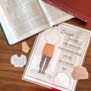 May include: A printable paper craft activity for kids featuring a paper doll representing Nebuchadnezzar's dream from the Bible. The dream is depicted as a large statue with different colored sections representing different empires. The statue has a gold head, silver chest and arms, bronze stomach and thighs, iron legs, and iron and clay feet. The statue is standing on a platform with a rock at the bottom. The text "Nebuchadnezzar's Dream Daniel 2" is at the top of the page.