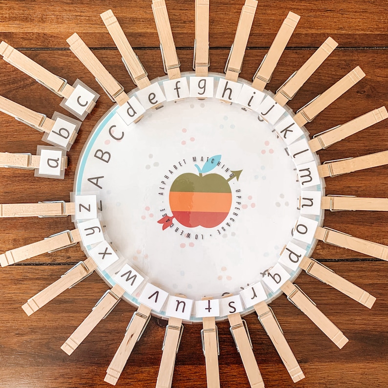 May include: Alphabet matching game with wooden pegs. The game features a circular design with uppercase and lowercase letters. Each peg holds a letter, and the centre has an apple graphic with the words "Alphabet Matching".