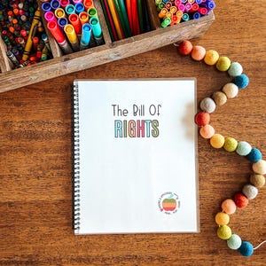 Puede incluir: Un cuaderno con espiral blanco con el texto "The Bill Of Rights" en letras coloridas. Un organizador de madera lleno de rotuladores y l&aacute;pices de colores est&aacute; en el fondo. Una guirnalda de bolas de fieltro de colores est&aacute; a la derecha.