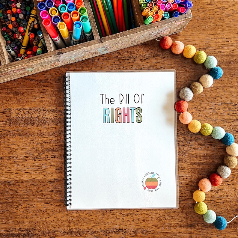 Puede incluir: Un cuaderno con espiral blanco con el texto "The Bill Of Rights" en letras coloridas. Un organizador de madera lleno de rotuladores y l&aacute;pices de colores est&aacute; en el fondo. Una guirnalda de bolas de fieltro de colores est&aacute; a la derecha.