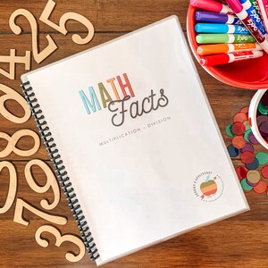 May include: A white binder with a colorful title that reads "MATH Facts Multiplication + Division". The binder is open and has a spiral binding. There are wooden numbers and colorful counters scattered around the binder.