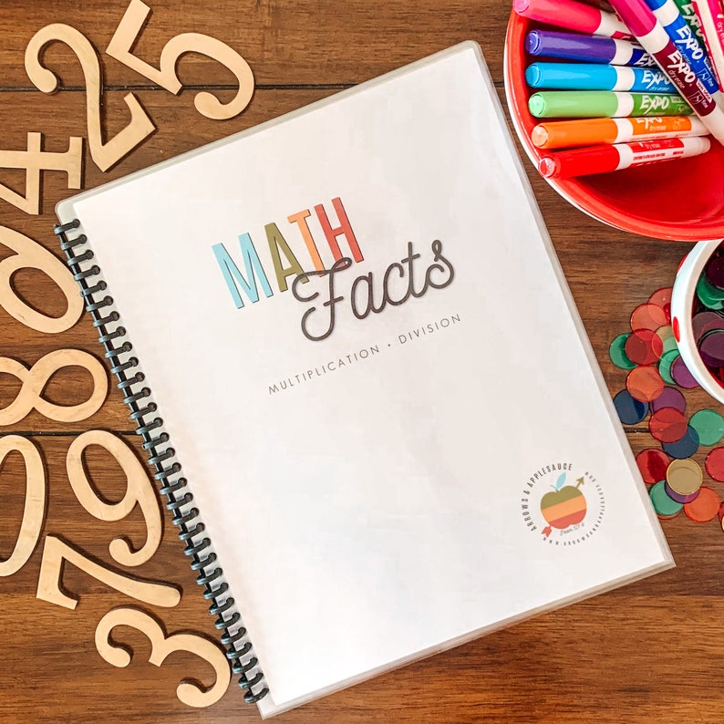 May include: A white binder with a colorful title that reads "MATH Facts Multiplication + Division". The binder is open and has a spiral binding. There are wooden numbers and colorful counters scattered around the binder.