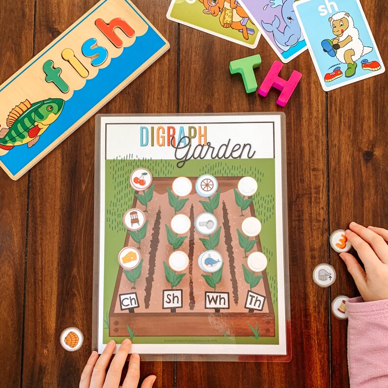 Puede incluir: Un juego de jard&iacute;n de d&iacute;grafos colorido con un marco de madera marr&oacute;n y hierba verde. El juego incluye una variedad de im&aacute;genes, como una cereza, una rueda, una silla, una lupa, un pez y un silbato. El juego tambi&eacute;n incluye un conjunto de fichas de letras que deletrean los d&iacute;grafos "ch", "sh", "wh" y "th".
